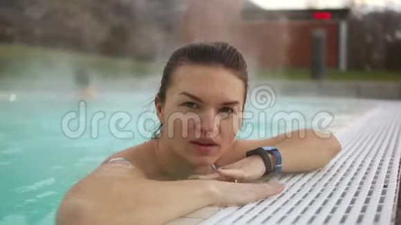 冬天在炎热的室外游泳池边缘附近美丽的黑发美女的亲密肖像蒸汽从水中升起女孩视频的预览图