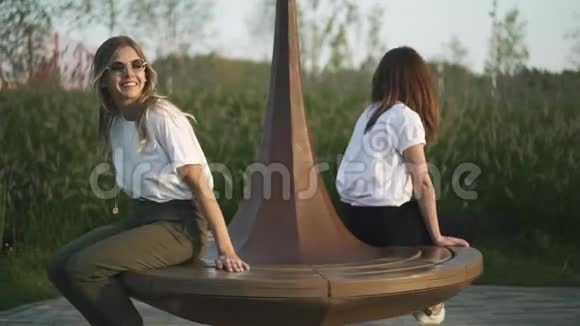 两个穿着t恤和太阳镜的年轻女孩在公园的长凳上旋转视频的预览图