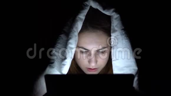 一个年轻的女人躺在毯子下面手里拿着一块药片黑暗中的女人用垫子现代电子平板电脑视频的预览图
