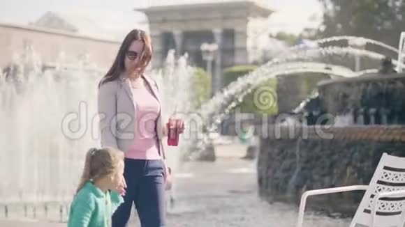 年轻的妈妈和小女儿在夏日公园的喷泉背景上散步视频的预览图
