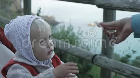 女人手给孩子食物穿着连帽衫和红色针织背心的女婴在山上坐婴儿车时吃火腿视频的预览图