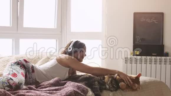 年轻漂亮的女人和她可爱的缅因州猫一起放松躺在床上用手机和听音乐视频的预览图