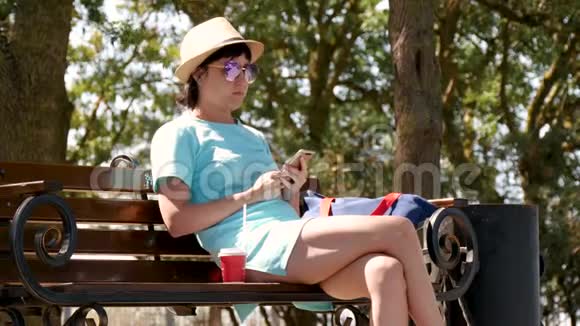 一个穿着帽子和裙子的黑发女孩坐在公园的长凳上用电话旁边的长凳上站着一杯咖啡视频的预览图