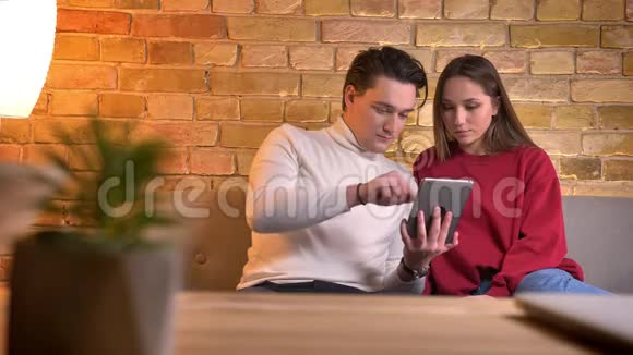 年轻的白种人夫妇坐在沙发上聚精会神地在家里观看平板电脑视频的预览图