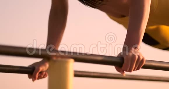 美丽苗条的女子运动员在日落时分穿着黑色上衣和黄色裤子在平行水平杆上做俯卧撑视频的预览图