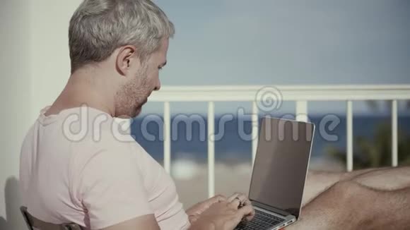 英俊的灰发男子在阳光明媚的海景露台上的笔记本电脑上工作视频的预览图