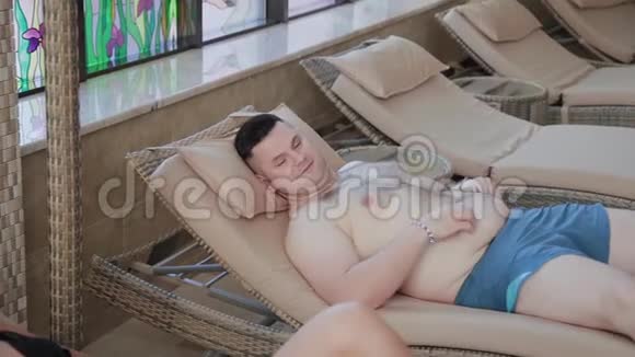 超重的男人和美丽的女人躺在被覆盖的游泳池旁的躺椅上视频的预览图