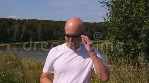阳光明媚的秃头在湖边的户外吹着太阳眼镜视频的预览图