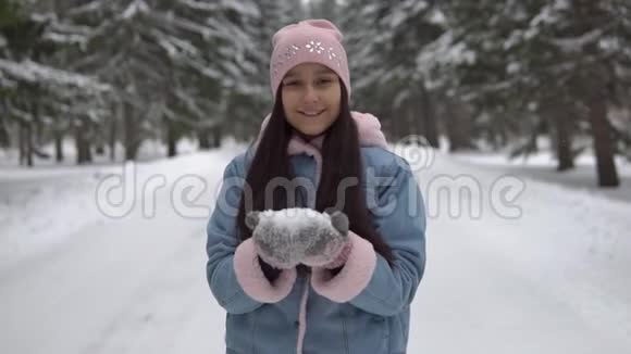 女孩站在冬天的森林里站在树的背景上用手吹雪微笑视频的预览图