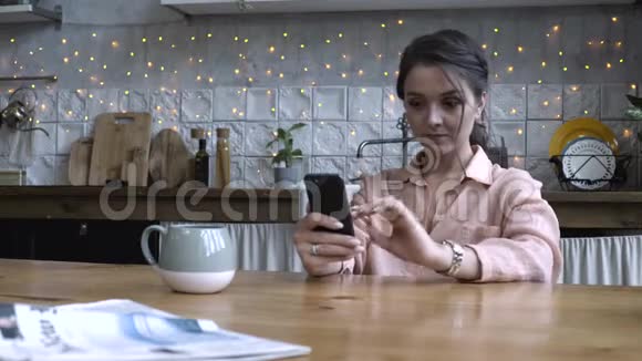 漂亮的年轻女人棕色头发穿着粉红色衬衫坐在厨房的木桌上用智能手机股票视频的预览图