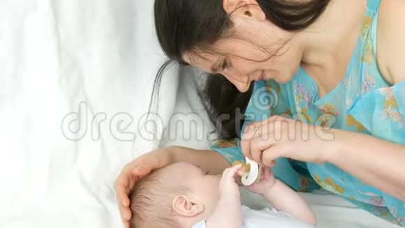 年轻漂亮的黑发妈妈躺在白色的床上和两个月大的新生儿玩耍视频的预览图