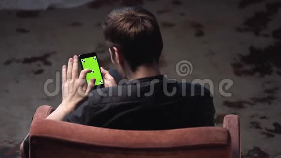在黑暗的房间里棕色头发的年轻人坐在椅子上敲着iPhone绿色屏幕库存数据视频的预览图