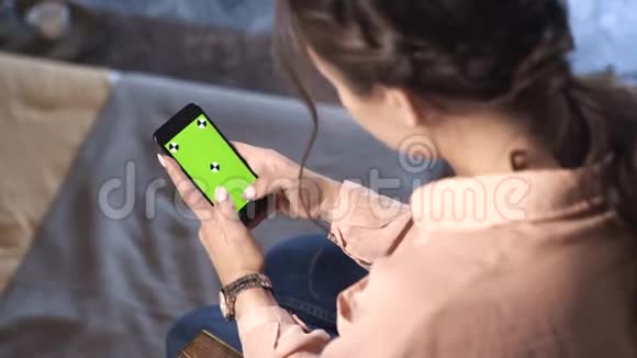 一个棕色头发的漂亮女人穿着粉色衬衫坐在家里在她的绿色屏幕上滑动着她的智能手机视频的预览图