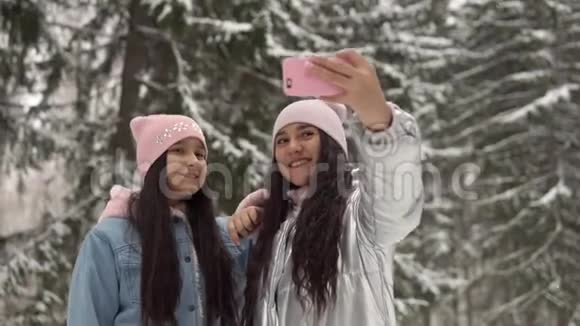 两个穿着冬天衣服的年轻女朋友用智能手机在冬天森林的背景下自拍4K视频的预览图