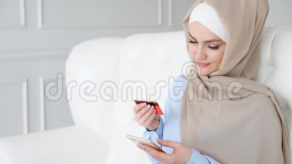 戴着头巾的穆斯林妇女正在用信用卡和智能手机在线购买视频的预览图