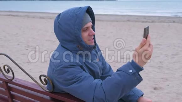 男子博主穿着蓝色羽绒服坐在沙滩的长椅上在手机上聊天视频视频的预览图