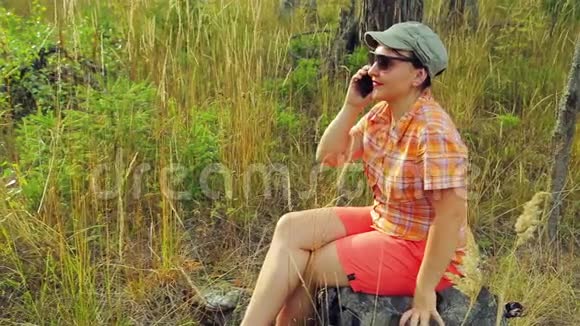 阳光明媚的一天戴着太阳镜和棒球帽的年轻女游客坐在一棵倒下的树上在电话里交谈视频的预览图