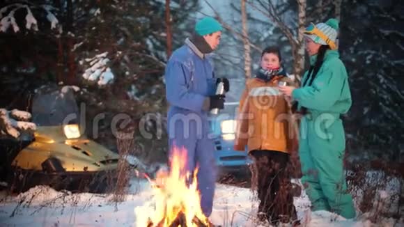 冬季森林年轻的一家人站在火边的树林里喝着泰莫斯的热饮视频的预览图