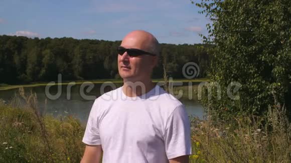 戴墨镜的秃头男子站在湖边的夏季森林里视频的预览图