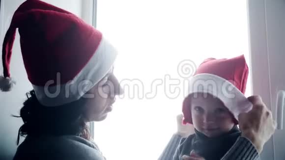快乐的家庭妈妈和宝宝在家玩白色窗户过圣诞节穿圣诞老人帽子的快乐男孩和女人视频的预览图