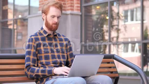 红胡子年轻人在笔记本电脑上工作坐在室外的长凳上视频的预览图