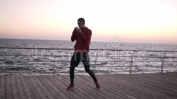 年轻的高个子男子在靠近大海的木地板上做拳击热身运动做附加步骤打孔热身运动视频的预览图