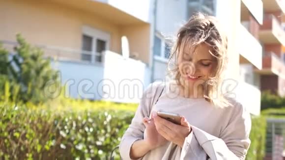年轻快乐的春天女人在一条城市街道上看着她的智能手机微笑阳光明媚的早晨视频的预览图