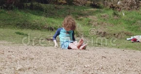 一个金发碧眼的小男孩穿着蓝色泳装坐在河滩上视频的预览图
