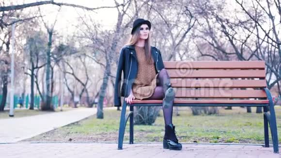 漂亮的年轻女子穿着时尚服装黑帽子脚踝靴和皮夹克独自坐在公园的长凳上视频的预览图