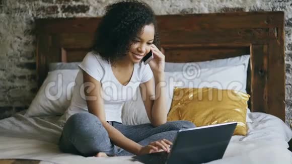 可爱的非裔美国女孩会说智能手机会用笔记本电脑坐在家里的床上视频的预览图