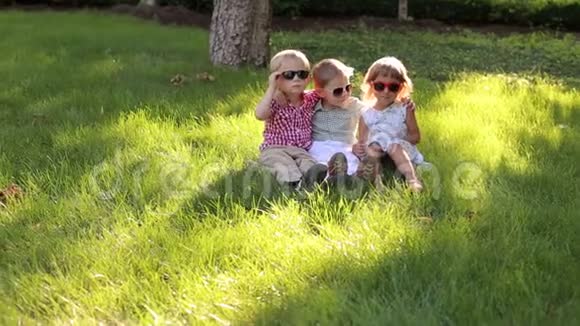 三个戴墨镜的快乐小朋友坐在公园的草地上视频的预览图