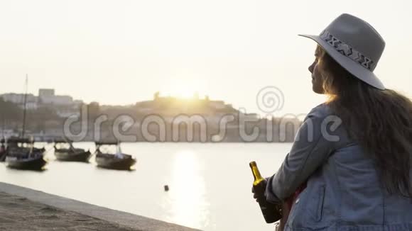 戴帽子的女士坐在河堤上喝一瓶酒视频的预览图