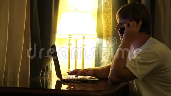一个年轻人坐在桌子旁在电脑上工作在手机上聊天视频的预览图