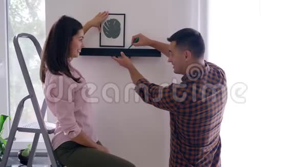 家庭装修新婚夫妇幸福地在墙上挂画和架子女孩子在公寓里亲吻男孩子视频的预览图