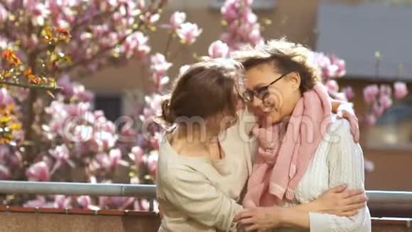 成熟的母亲和成年的女儿拥抱站在阳台上背景是玉兰花的开花树明亮明亮视频的预览图