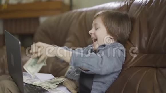 六岁的小男孩坐在皮沙发上腿上放着笔记本电脑把钱扔到空中视频的预览图