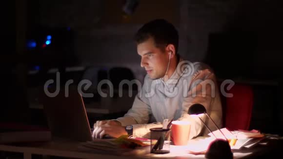 可爱的白种人休闲男性正在用耳机听音乐坐着移动用笔记本电脑打字视频的预览图