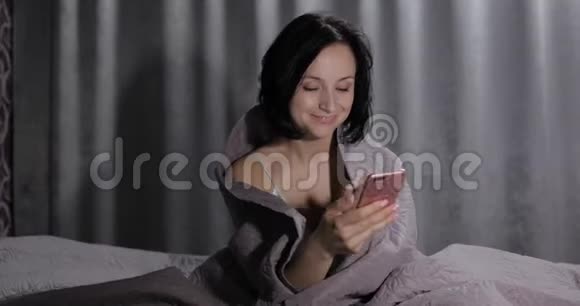 女人坐在毯子下面的床上喜欢在智能手机上与朋友视频聊天视频的预览图