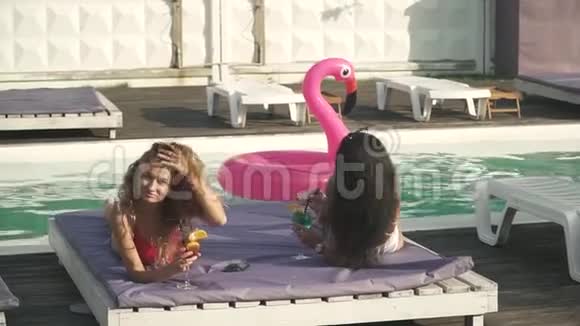 在游泳池附近的休息室里有两个漂亮的女孩穿着漂亮的泳装梳着鸡尾酒谈得很愉快视频的预览图