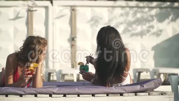 在游泳池附近的休息室里有两个漂亮的女孩穿着漂亮的泳装梳着鸡尾酒谈得很愉快视频的预览图