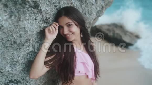 微笑美丽女孩的特写肖像在海滩上的岩石附近摆着大波浪慢动作视频的预览图