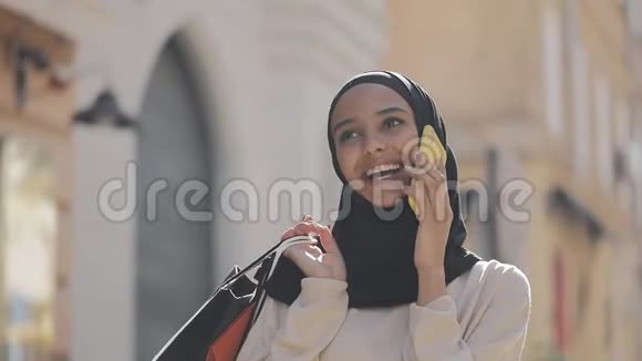 幸福的年轻穆斯林妇女戴着头巾在智能手机上说话手里拿着购物袋走在街上视频的预览图