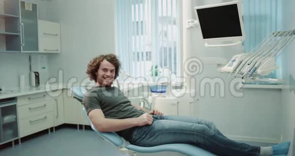 他坐在一家牙科诊所里一位可爱的有魅力的卷发笑容满面的男士预约了他的牙医视频的预览图