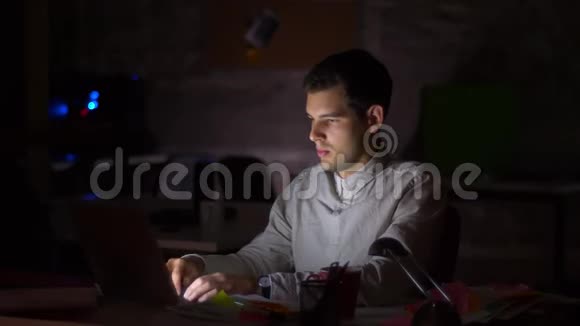 英俊的白种人男性坐在桌面上在笔记本电脑上工作在黑暗中为屏幕发光视频的预览图