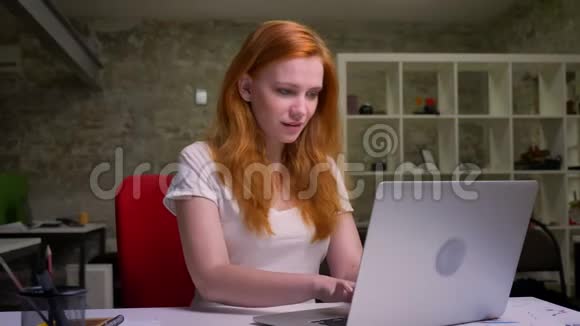可爱的微笑的白种人女孩正在笔记本电脑里打字而她正坐在桌面上的砖光办公室里脸很自然视频的预览图
