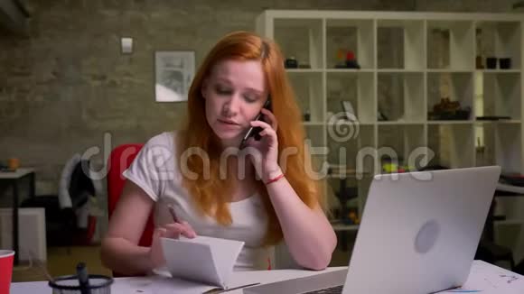 辛勤工作的白种人姜家女商人正在电话交谈并在笔记本电脑办公室插图附近查看网站视频的预览图