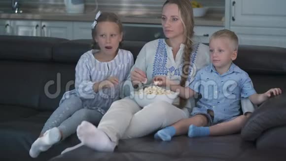 妈妈和两个孩子坐在沙发上吃爆米花看电视视频的预览图