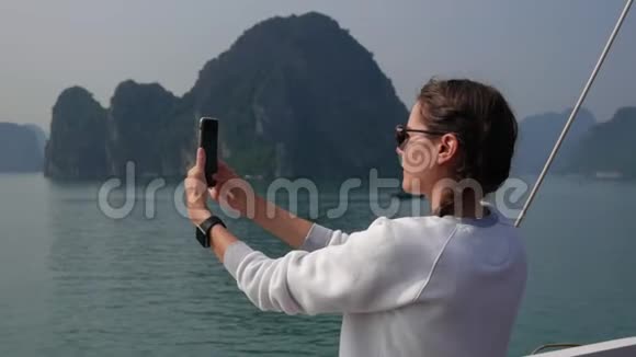 年轻女子穿着白色毛衣和太阳镜带着智能手表站在智能手机上拍摄海湾的照片视频的预览图