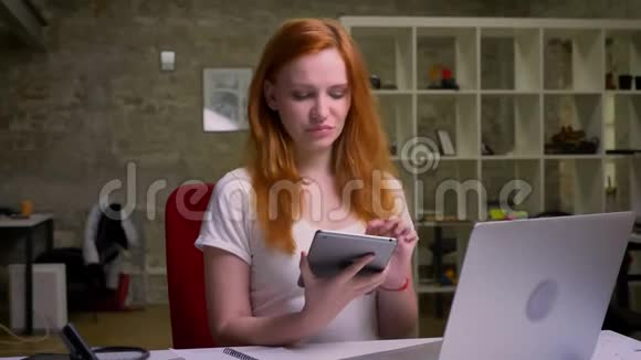 可爱快乐的白种人红发女孩正孤立地坐在办公室里手里拿着绿色屏幕的平板电脑视频的预览图