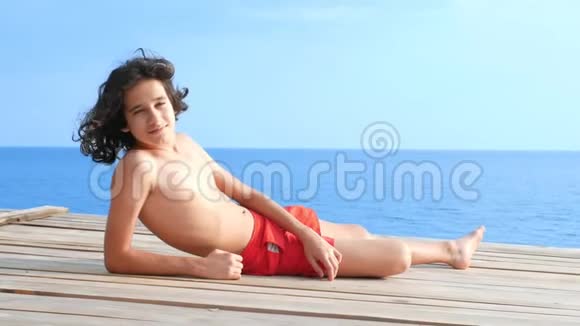 一个长着卷曲黑发的英俊少年躺在海面上的一个木制露台上暑假概念学校视频的预览图